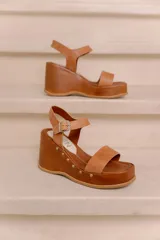Sandalias de plataforma y taco alto confeccionadas en cuero vacuno marrón, con tira ancha en el empeine, tira ajustable con hebilla en el tobillo y detalles de tachas decorativas en la base.