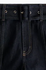 Pantalón de jean de tiro alto con pierna amplia y corte holgado. Confeccionado en denim rígido de color azul oscuro, presenta costuras decorativas verticales en el frente, bolsillos laterales y un cinturón integrado del mismo material con hebilla.