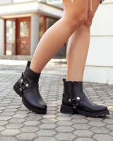 Bota de caña alta confeccionada en cuero negro, con suela track de goma. Presenta un diseño versátil con dos hebillas ajustables en la parte superior de la caña y una correa decorativa con tachas metálicas y argollas en la zona del tobillo.