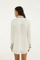 Camisa blanca de manga larga y calce relajado, confeccionada en tejido liviano de textura arrugada. Presenta cuello camisero, botonera frontal y detalles de ribete en zigzag marrón oscuro en cuello, puños y dobladillo.