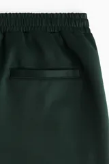 Pantalón deportivo de corte relajado en tejido efecto neopreno, color verde oscuro con franjas laterales en contraste color crema. Presenta cintura elástica con cordón ajustable, bolsillos laterales y traseros, y cierre oculto en el bajo.