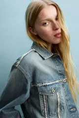 Chaqueta de jean estilo crop, con cuello solapa, manga larga y cierre frontal con botones metálicos. Presenta bolsillos de plastrón con solapa en el pecho y detalles de costuras deshilachadas en el frente.