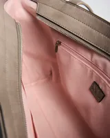 Cartera tipo tote de símil cuero color taupe, con doble asa de hombro, detalles de tachas plateadas en los laterales frontales y placa metálica con logo grabado de la marca XL Extra Large en el centro.
