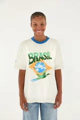 Camiseta blanca de algodón con cuello redondo azul, manga corta y gran estampado frontal con la palabra BRASIL, la bandera de Brasil y un pájaro verde en vuelo.