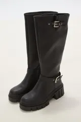 Bota de caña alta color negro, confeccionada en eco cuero, con suela track de goma y detalle de hebilla metálica en el tobillo y en la parte superior de la caña.