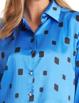 Camisa de satén de corte holgado con cuello camisero y cierre frontal de botones metálicos. Presenta un estampado de cuadrados negros sobre fondo azul vibrante, mangas largas con puños abotonados y un acabado brillante característico del satén.