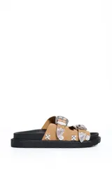 Sandalia plana color beige con dos tiras anchas decoradas con hebillas grandes y apliques metálicos plateados de estilo western, sobre suela negra.