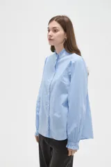 Camisa celeste de manga larga con cuello alto y volados, detalles de frunces en hombros y puños, y cierre frontal con botones.