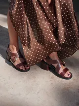 Sandalias de cuero color marrón con tiras anchas cruzadas y hebilla metálica decorativa en forma de estribo. Presentan taco cuadrado de altura media y suela de goma.