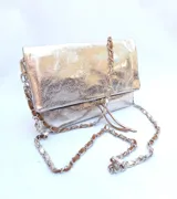 Cartera plateada tipo bandolera con solapa y cierre magnético. Tiene una cadena corta y una cadena larga.