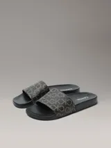 Ojotas Calvin Klein con diseño de monograma de la marca en color beige sobre fondo negro. Presentan una suela anatómica contorneada para mayor confort y una tira superior de material sintético.