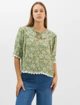 Blusa de gasa estampada con motivos botánicos en color verde, con escote redondo, abertura frontal con lazos de ajuste y mangas cortas. Presenta detalles de encaje en el escote, mangas y ruedo.