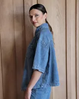 Sobrecamisa de denim soft en tono azul medio, con corte oversize, mangas cortas, cuello clásico y cierre frontal con botones metálicos. Presenta dos bolsillos frontales con solapa y un ruedo redondeado.