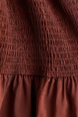 Vestido corto marrón de corte entallado, con diseño fruncido tipo nido de abeja en todo el cuerpo, escote cuadrado con hombros descubiertos y volados anchos en el escote y el ruedo.