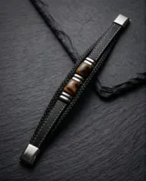 Pulsera de diseño masculino compuesta por una banda de cuero sintético trenzado en color negro, con detalles centrales de acero y piedras naturales ojo de tigre.