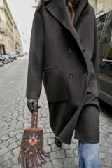 Abrigo largo cruzado color gris oscuro, confeccionado con lana. Presenta cuello solapa, manga larga, bolsillos delanteros de plastrón y cierre frontal con botones.