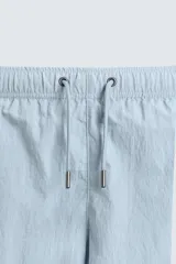 Bañador de hombre de largo medio, color celeste claro con rayas blancas horizontales. Presenta cintura elástica ajustable con cordón, bolsillos frontales y bolsillos traseros de plastrón. Incluye forro interior.