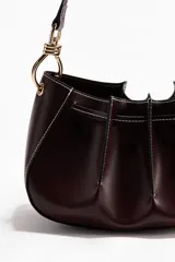 Cartera de hombro de tejido revestido color bordó, con pliegues cosidos, detalles de pespuntes en contraste, cordón ajustable superior y asa larga con herrajes metálicos dorados.
