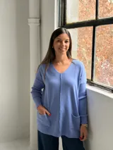 Sweater de punto color azul lavanda, con escote en V, mangas largas con puños acanalados y dos bolsillos frontales tipo parche. Presenta una costura central vertical visible.