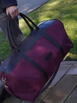 Bolso de viaje tipo duffel de lona negra con detalles en cuero tostado, asas de mano y correa ajustable para el hombro.