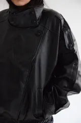 Chaqueta de cuero sintético color negro, con diseño de corte oversize, cuello alto con cierre de botones a presión, hombreras marcadas y ruedo elástico.