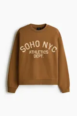 Buzo marrón de algodón con cuello redondo, hombros caídos y diseño estampado en el frente con la inscripción 'SOHO NYC ATHLETICS DEPT.' en color beige. Presenta puños y bajo de canalé elástico.