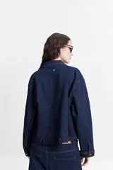 Campera de jean de mezclilla oscura, corte relajado, con cuello y bolsillos de parche con tapa y botón en el pecho. Presenta costuras en tono naranja/marrón claro.