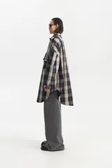 Camisa de largo extendido, estilo camisaco, con patrón de cuadros escoceses en tonos negro, blanco crema y gris lavanda. Presenta cuello camisero, cierre frontal con botones y dos bolsillos de parche con tapa en el pecho. El corte es holgado y la parte inferior es redondeada.