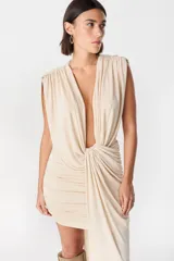 Vestido corto color beige con escote profundo en V, diseño drapeado en la cintura y hombreras marcadas. Presenta una falda ajustada con frunces laterales y una caída asimétrica lateral.