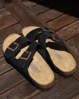 Sandalias tipo zueco marca Rivaro, confeccionadas en cuero gamuzado negro con dos tiras ajustables con hebillas metálicas. Presentan una plantilla anatómica de corcho y látex, y suela de goma antideslizante.