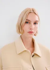 Chaqueta de corte recto color beige, con cuello camisero, cierre frontal de botones oscuros y cuatro bolsillos con solapa en el frente.