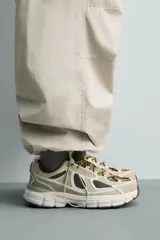 Zapatilla deportiva tipo running con diseño de suela voluminosa e irregular. Presenta una combinación de piezas en tonos beige y crema, con paneles de malla y cordones jaspeados.