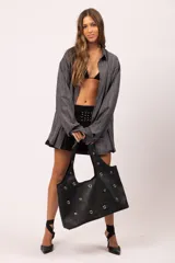 Cartera tipo tote de eco cuero negro con textura granulada, decorada con múltiples ojalillos metálicos plateados distribuidos en el frente y las asas. Cuenta con cierre de botón y forro interior con bolsillo.