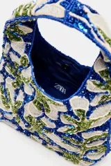 Bolso de mano mini con diseño floral bordado en abalorios y lentejuelas en tonos azul, verde y blanco. Cuenta con asa integrada, forro interior con bolsillo y cierre mediante botón imantado.