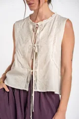Blusa sin mangas de color beige, confeccionada en tela ligera con detalles de bordados florales frontales, encajes y cierre delantero con tiras para anudar.