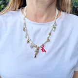 Collar corto hecho con cinta de tela blanca y dijes dorados, rojos y nacarados con forma de pez, corazón, coral, flor y cuentas.