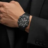 Reloj de pulsera analógico con cronógrafo, caja redonda de metal negro y correa de caucho negra. Presenta esfera texturizada con marcadores luminosos y detalles en rojo.
