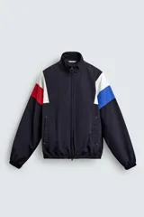 Chaqueta técnica de corte relajado con cuello subido y cierre frontal de cremallera bidireccional. Presenta un diseño color block en tonos azul marino, blanco, rojo y azul eléctrico. Incluye bolsillos frontales de vivo, puños y ruedo elásticos, y forro interior de red.