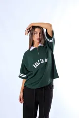 Chomba de piqué color verde con cuello polo blanco y cierre frontal parcial. Presenta un diseño holgado con mangas cortas que incluyen rayas blancas en los puños y un aplique frontal con el texto 'HOLE IN ONE' en color blanco.