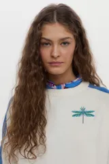 Sweater de punto color blanco con mangas raglán en azul y detalles en verde en los puños, cuello redondo y bordado de libélula en el pecho.