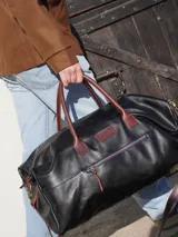 Bolso de viaje tipo weekender confeccionado en cuero negro, con manijas y correa ajustable en cuero color bordó. Presenta cierre metálico frontal con detalle en azul, bolsillo externo con cierre y parche de marca grabado en el frente.