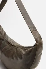 Bolso de hombro tipo media luna, fabricado en piel craquelada color marrón oscuro. Tiene un compartimento principal con cierre de cremallera y un bolsillo interior también con cremallera.