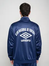 Conjunto deportivo Umbro del Club Nacional de Football, compuesto por una campera con cierre frontal y pantalón a juego. La campera es de color azul con detalles en rojo en los hombros y líneas blancas, presentando el logo de la marca y el escudo del club en el pecho. El conjunto está confeccionado en tejido sintético con acabado satinado.