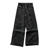 Pantalon baggy-flared de denim negro con efecto gastado, diseño desmontable con cierres horizontales en las piernas. Presenta múltiples parches de refuerzo, apliques metálicos, ojales decorativos y correas ajustables en los tobillos. Incluye un cinturón textil con tachas.