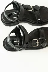 Sandalias negras de estilo chunky con plataforma dentada. Presentan tiras ajustables con cierre de velcro y hebillas metálicas, combinando texturas de cuero granulado y detalles en charol.