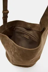 Bolso tipo saca de diseño asimétrico confeccionado en piel de ante. Cuenta con un asa bandolera ajustable, cierre mediante imán y un bolsillo interior con cremallera.
