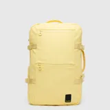 Mochila de viaje color amarillo pastel, con diseño rectangular y compartimentos frontales con cierre. Incluye correas laterales ajustables y asa lateral para transporte tipo maletín. Cuenta con etiqueta de la marca en la parte inferior.