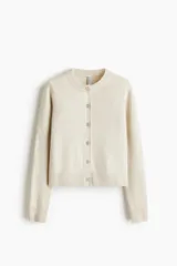 Saco entallado de punto fino en color beige, con cuello redondo, cierre frontal de botones y detalle de corazón bordado en el frente. Presenta terminaciones acanaladas en cuello, puños y bajo.