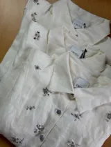 Camisa de lino color crudo con cuello clásico y cierre frontal de botones. Presenta bordados artesanales florales distribuidos por toda la prenda, realizados a mano.