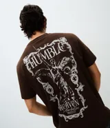 Remera de algodón color marrón con cuello redondo y mangas cortas. Presenta un diseño gráfico de estilo barroco con una figura alada y la inscripción 'Humble' en la espalda.